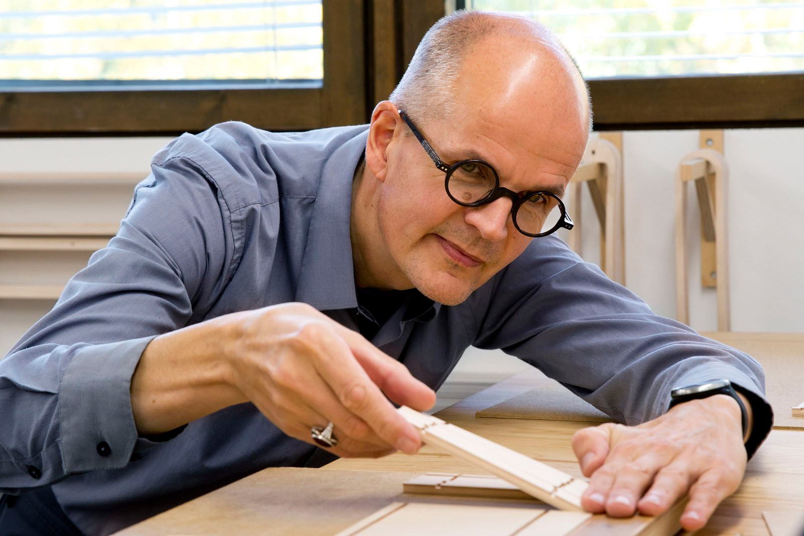 A smiling man wearing black round glasses and a blue shirt, focused on setting small slats on the jig.