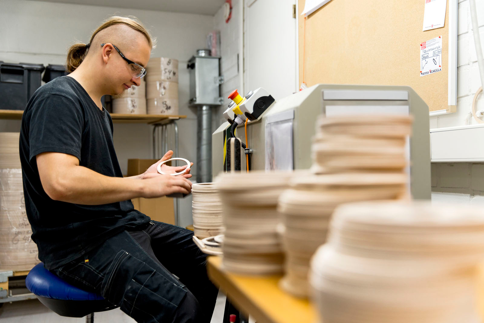 A male employee sitting on a chair on the left, sanding the wooden rings, which are piled up on a table on the right.