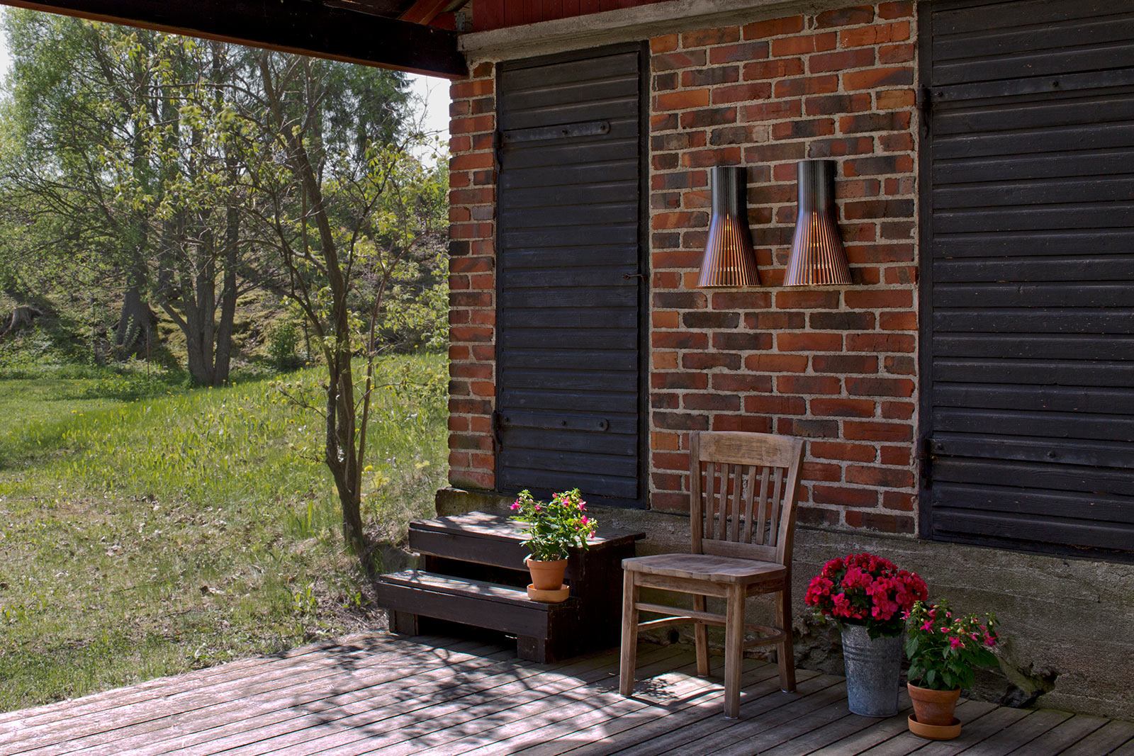 A covered patio with flowers and a chair in front of a brick wall. Two Secto Small wall lamps between black doors.