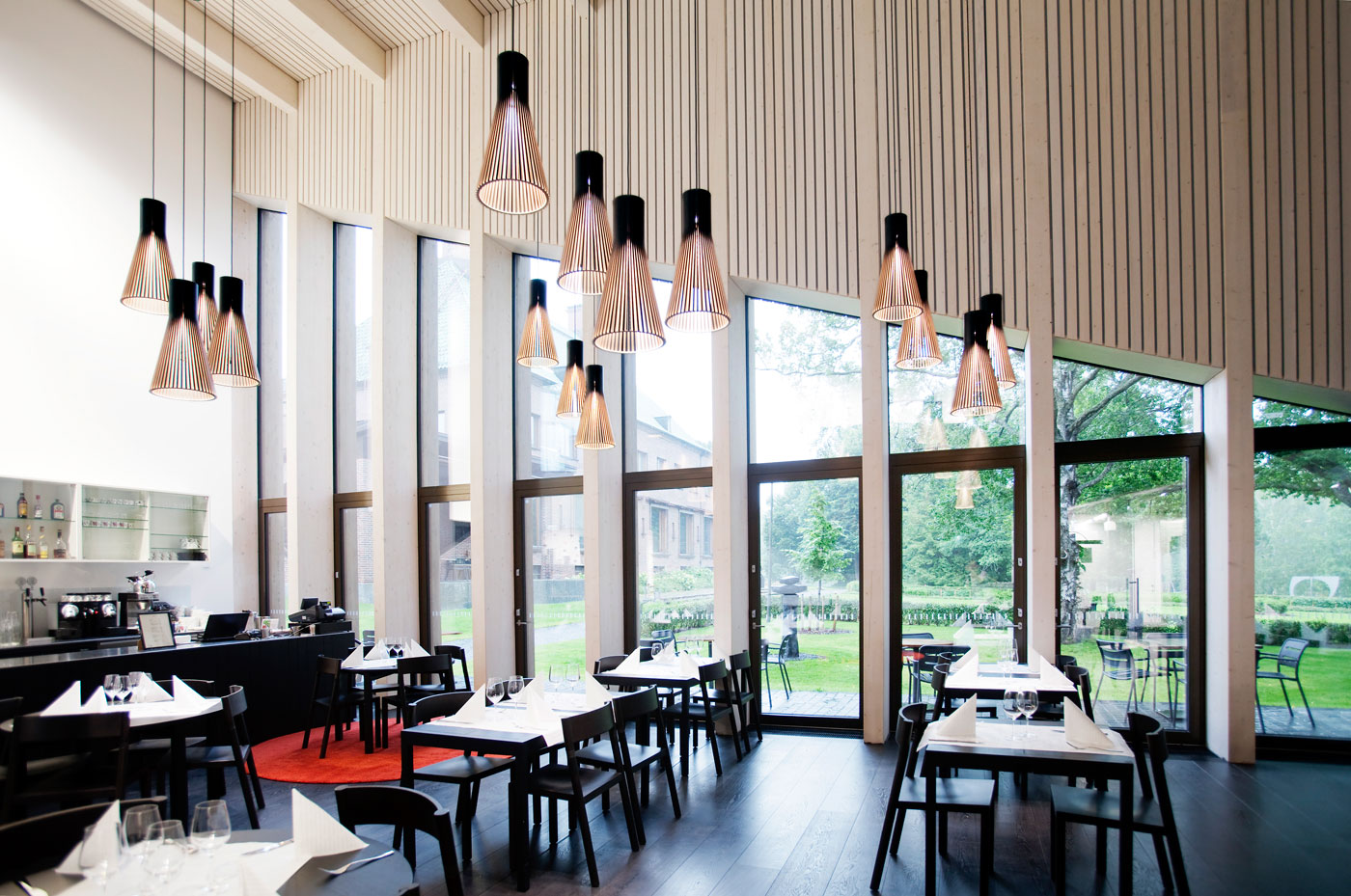 A dining hall with a bar on the left side and large windows towards a garden. Secto pendant lamps hanging above tables.