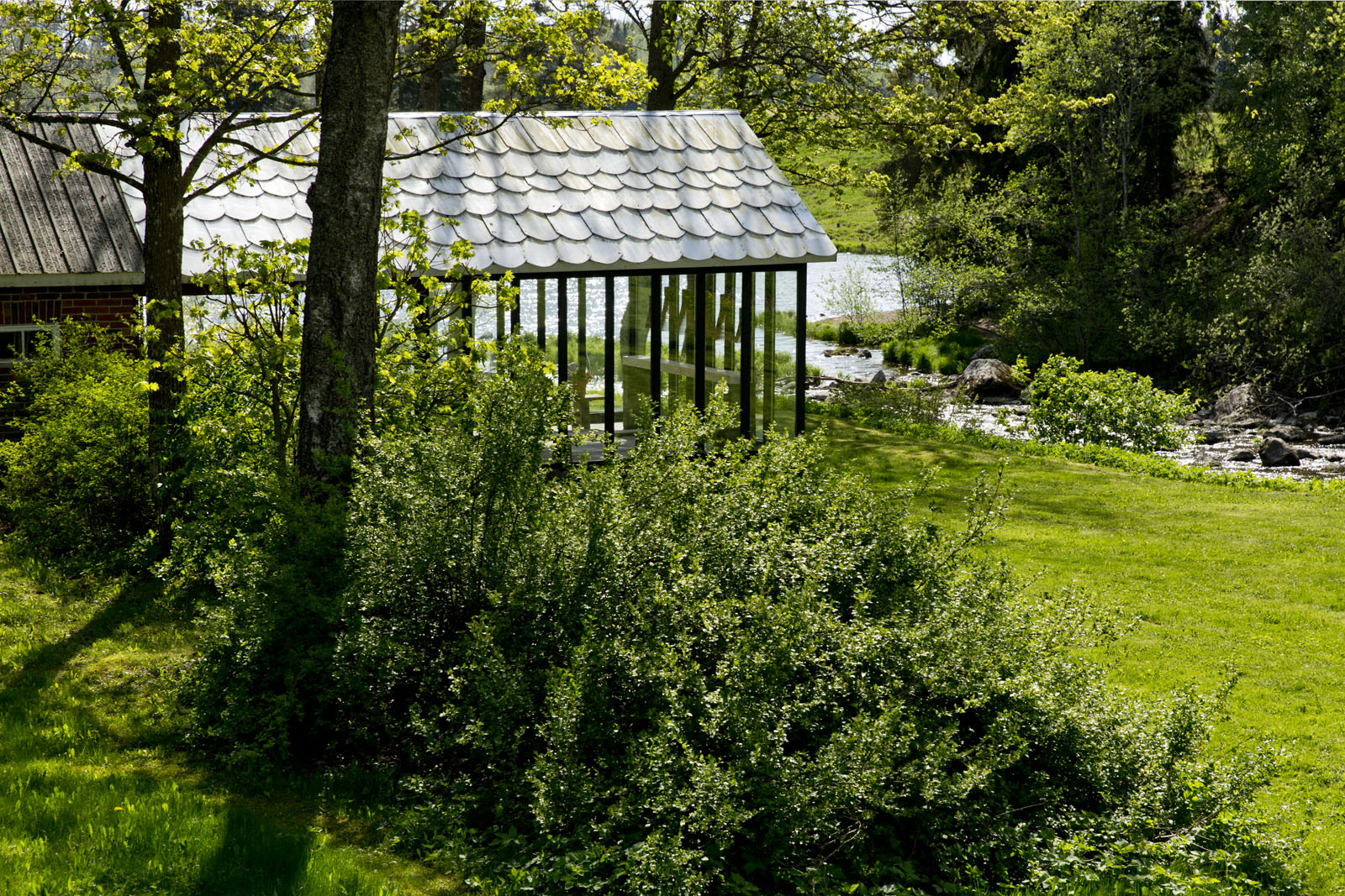Une maison en briques avec un porche en verre sur la gauche, une petite rivière se jette dans un lac depuis la droite à travers le jardin.