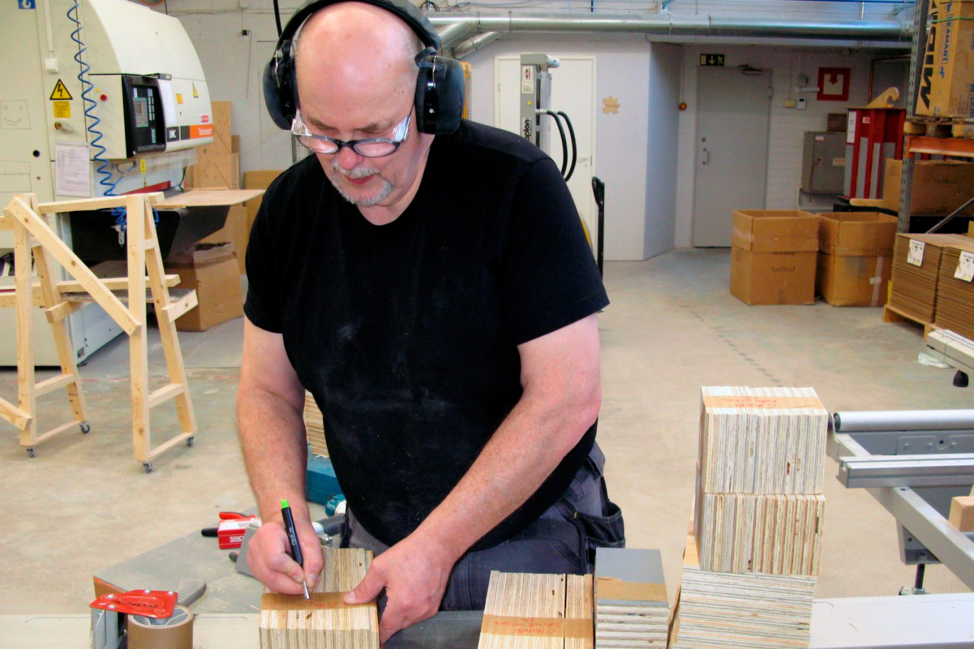 Un homme dessine des marques sur des blocs de bois. Il porte une protection auditive et des lunettes de sécurité.